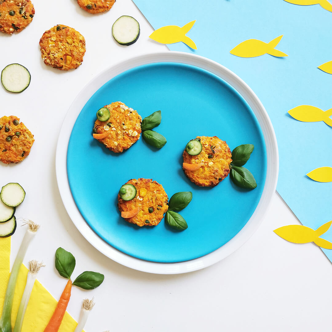 3 Baby Salmon Fish Cakes on a plate. The fish cakes have cucumber and peas for eyes a carrot mouth and basil leaves for tails