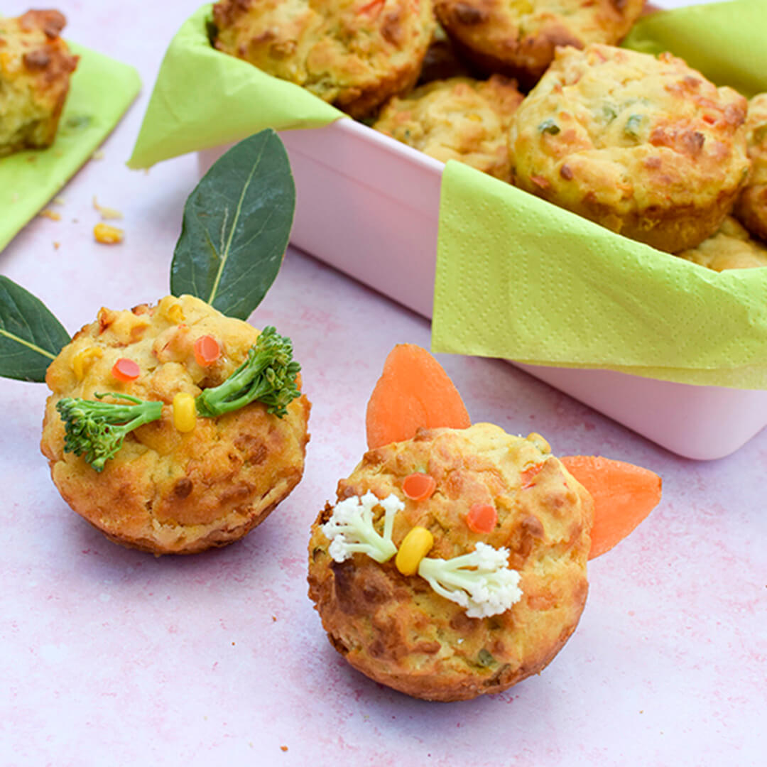 A container of Savoury Vegetable Muffins for Babies & Toddlers next to two savoury vegetable muffins - one of them has a sweetcorn nose with small broccoli floret whiskers and herb leaves for ears, the other has a sweetcorn nose, small cauliflower floret whiskers and carrot slices for ears