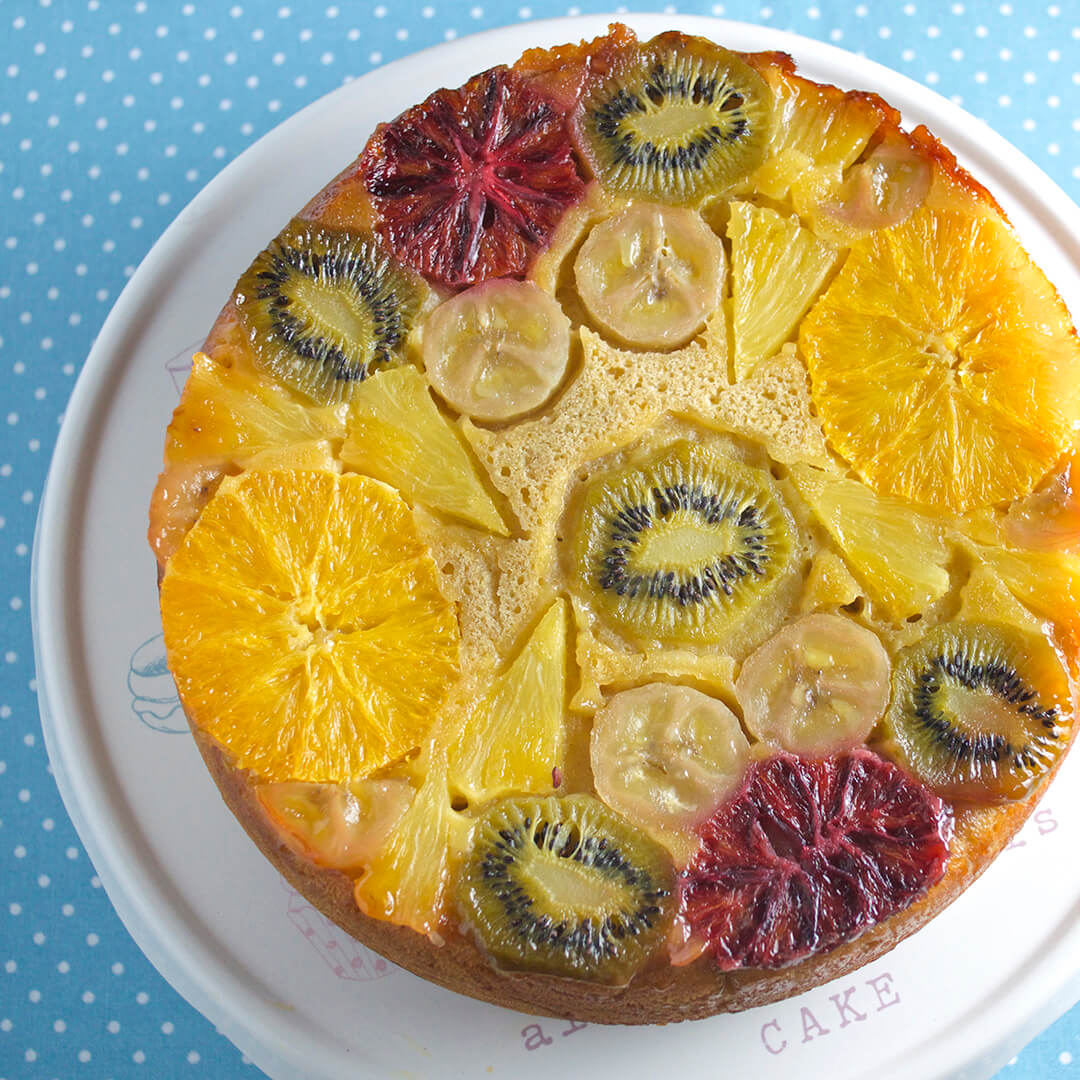 Tropical upside down cake on a cake stand. The bottom of the cake has kiwi, orange, blood orange, banana and pineapple chunks