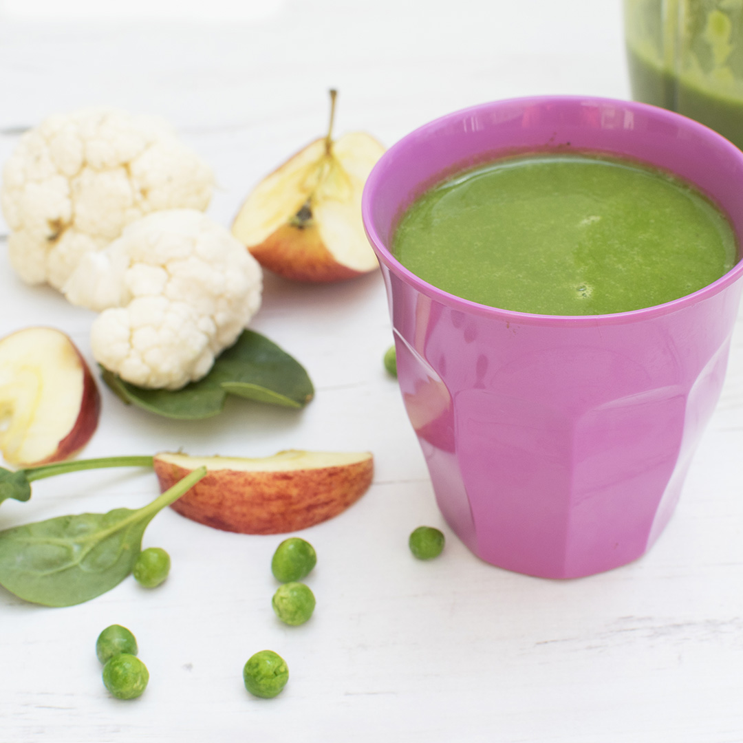 Green smoothie in a purple cup next to some cauliflower florets, peas, spinach leaves and a sliced apple