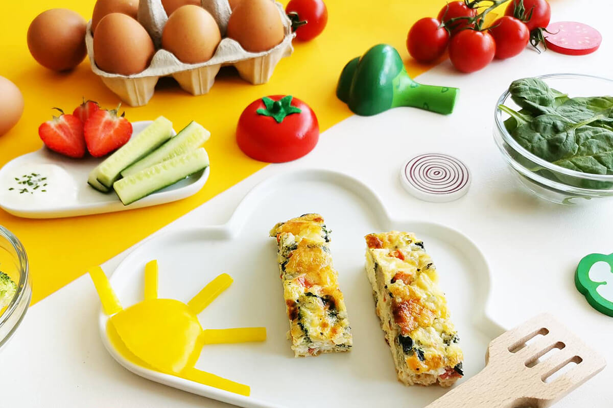 A serving of 2 omelette fingers on a plate next to a case of 6 eggs, some tomatoes, spinach, cucumber sticks and strawberries