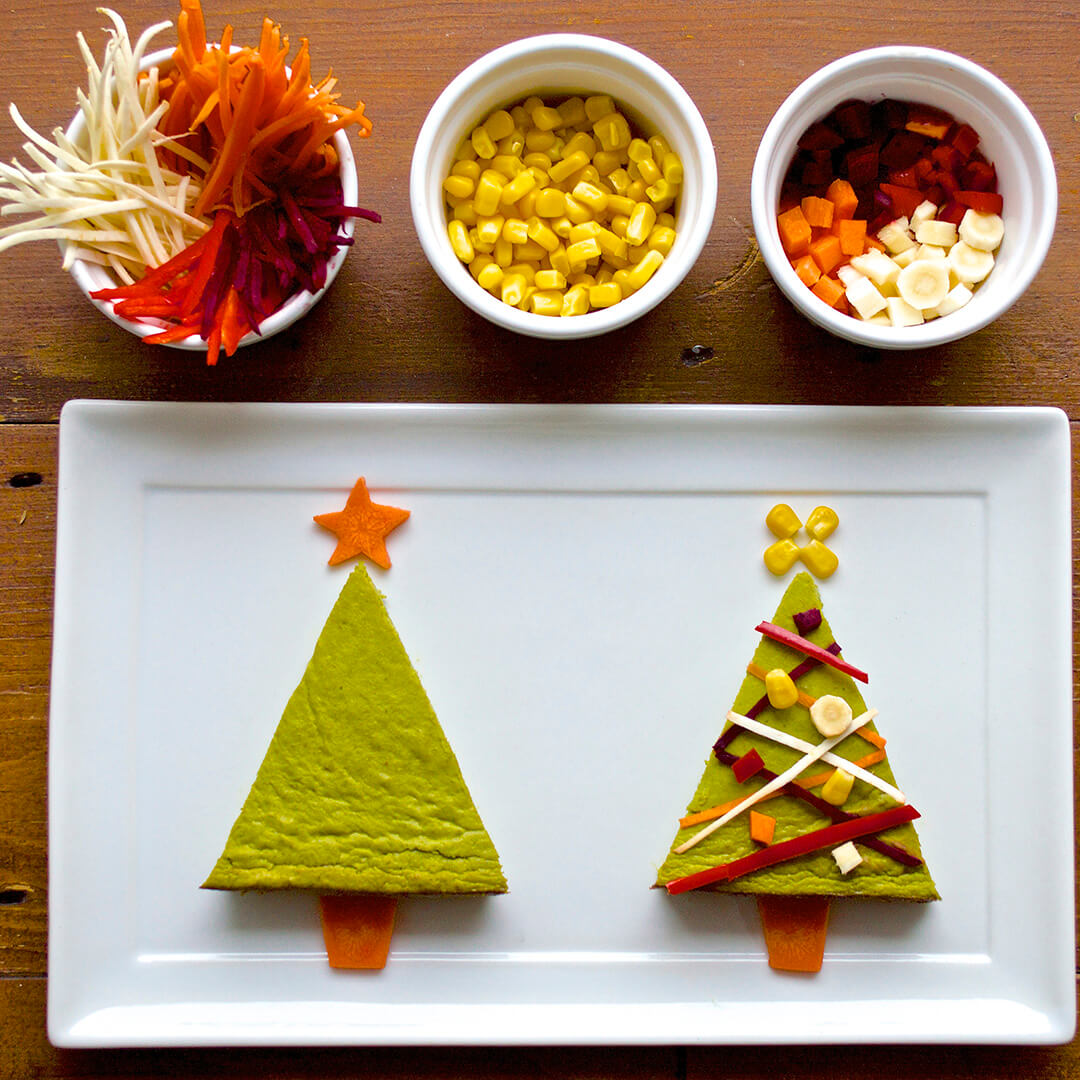 2 Christmas Tree tarts decorated with vegetables next to bowls sweetcorn, diced cooked sweet potato and other vegetables, and shredded cabbage, carrot and parsnip