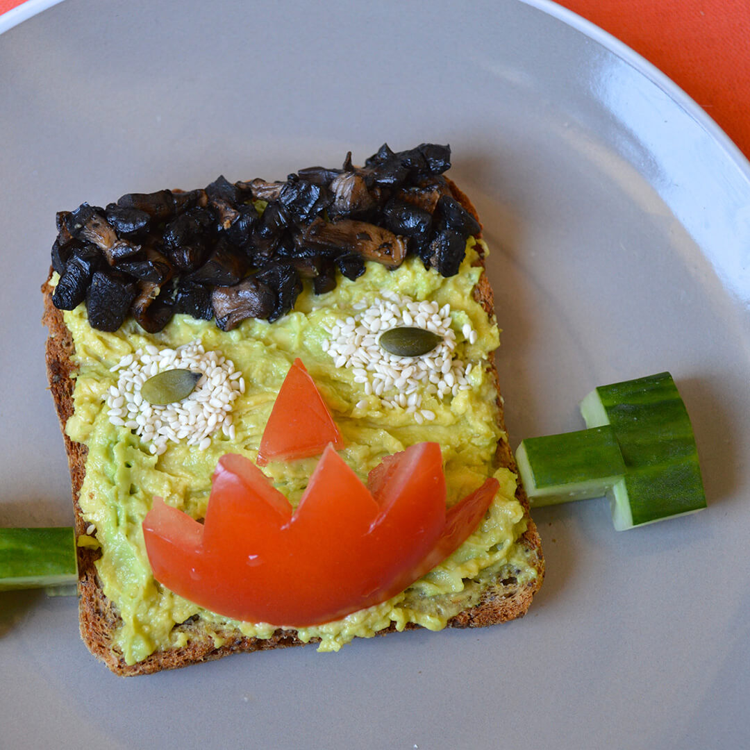 Halloween Frankenstein Toast with avocado skin, mushroom hair, sesame and pumpkin seed eyes, tomato nose and mouth and cucumber ears