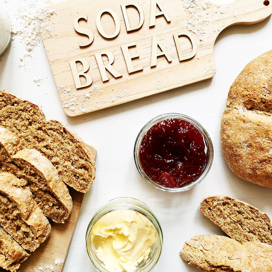 A small load of soda bread next to some sliced soda bread and small jars of jam and butter/cream