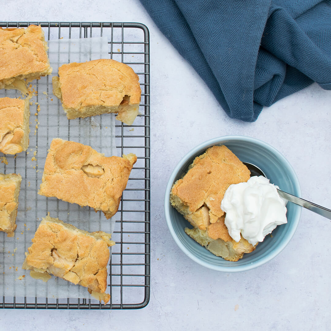 A slice of apple tray bake in a bowl with cream, next to a cooling rack of slices of apple traybake