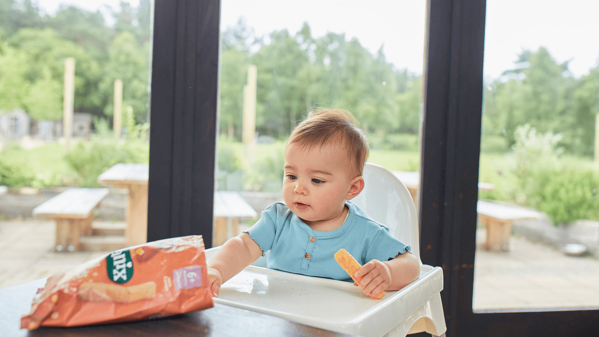 Baby on a highchair eating an Organix Weaning Wand and reaching out for the bag of Organix Weaning Wands