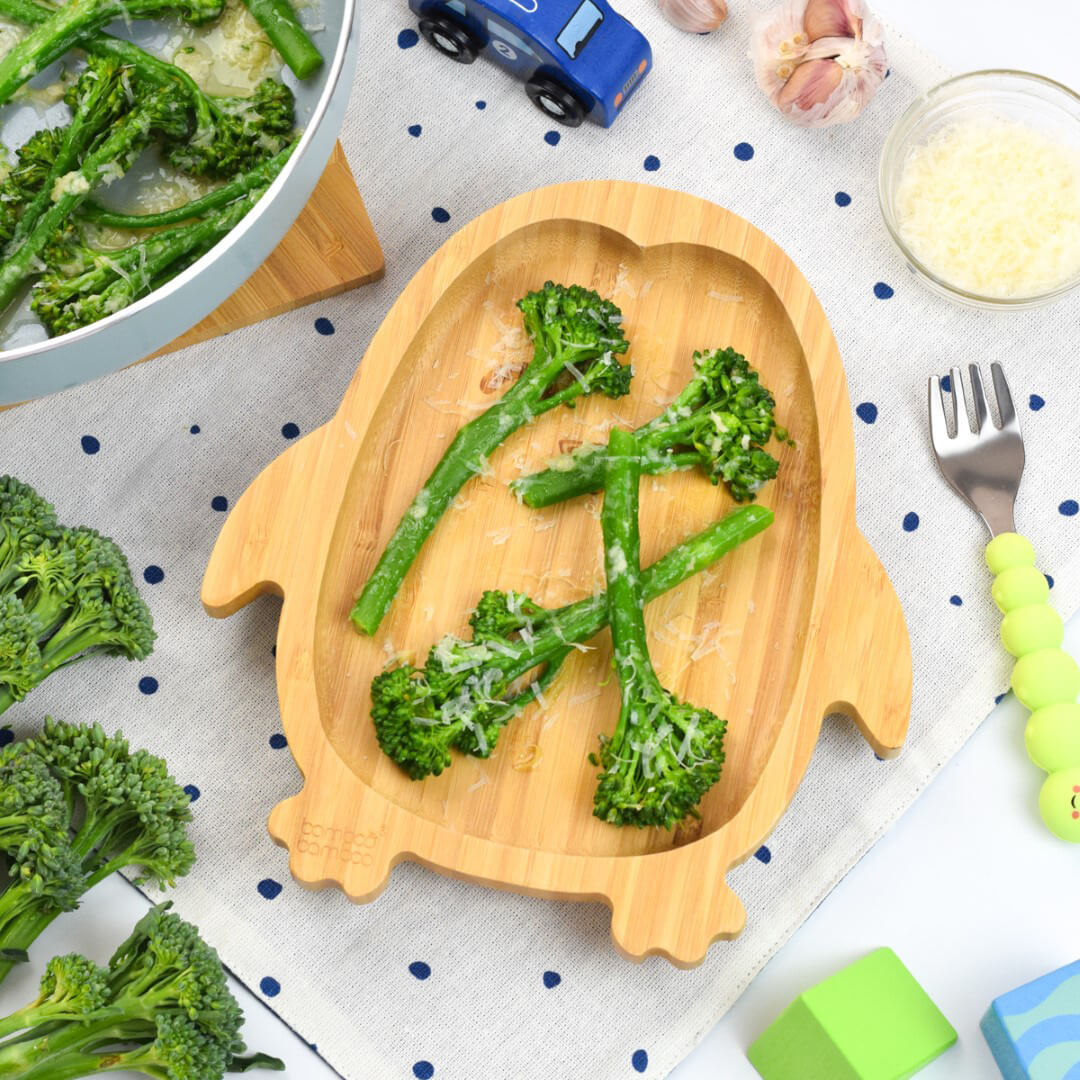 A serving of Parmesan broccoli next to some tenderstem broccoli florets, a small bowl of Parmesan cheese and a garlic clove