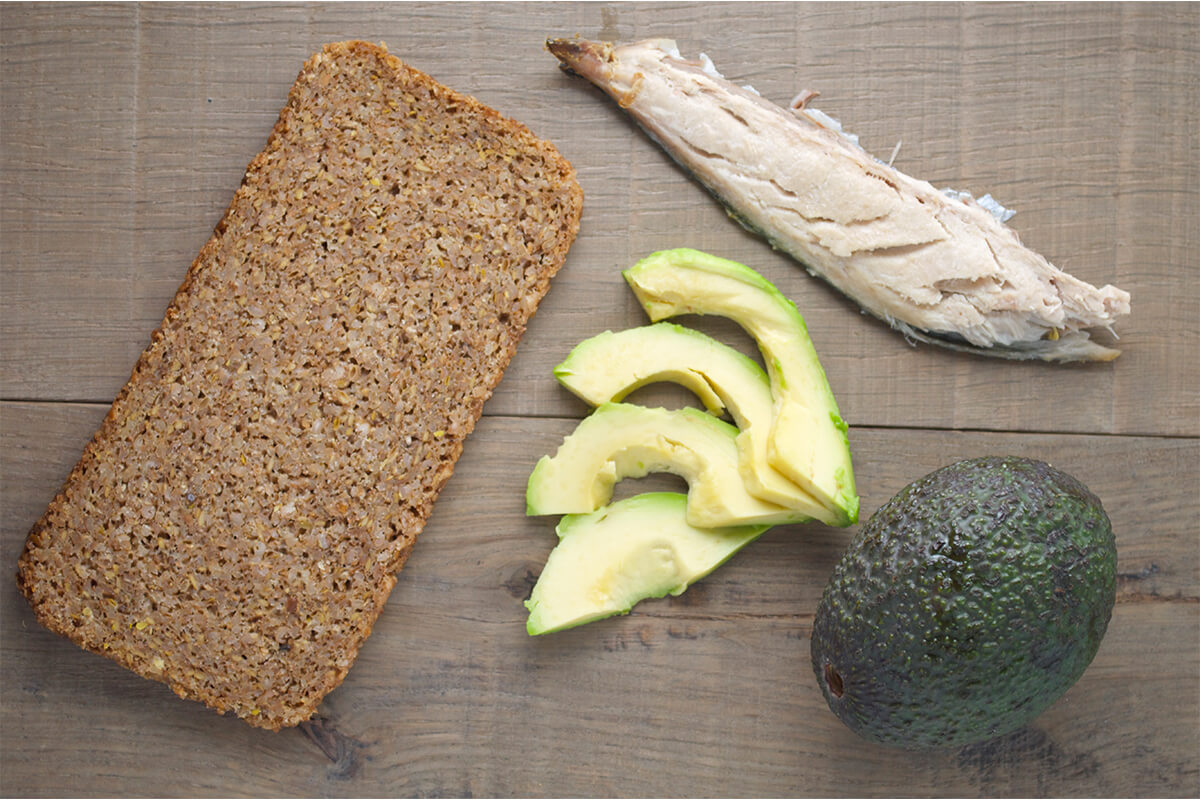 A slice of rye bread next to a filet of mackerel, a whole avocado and some sliced avocado