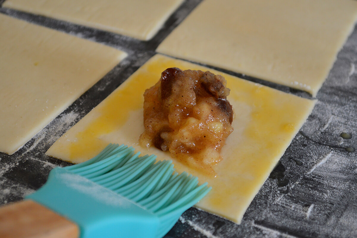 4 pastry squares on a floured surface, one with apple mixture in the middle while beaten egg is brushed around the edge