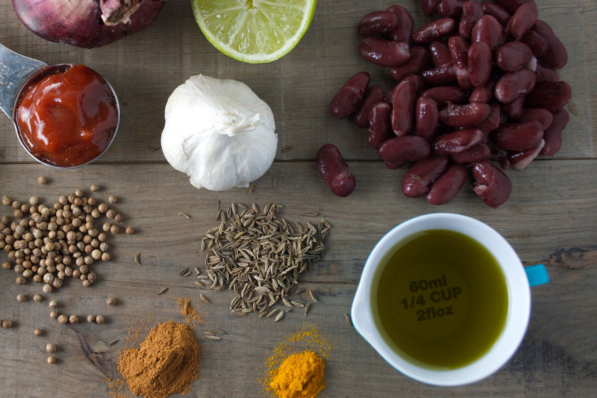 A table with a red onion, halved lime, red kidney beans, garlic clove, tomato puree, cumin, coriander, cinnamon and olive oil