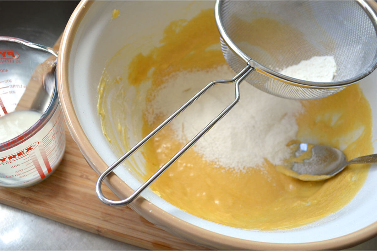 A mixing bowl with cake batter and flour being sieved into it, next to a glass jar of buttermilk