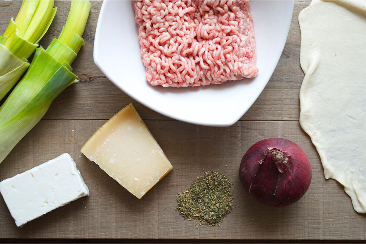 A table with some leeks, a white bowl of pork mince, rolled out pastry, red onion, dried herbs and small blocks of feta and parmesan