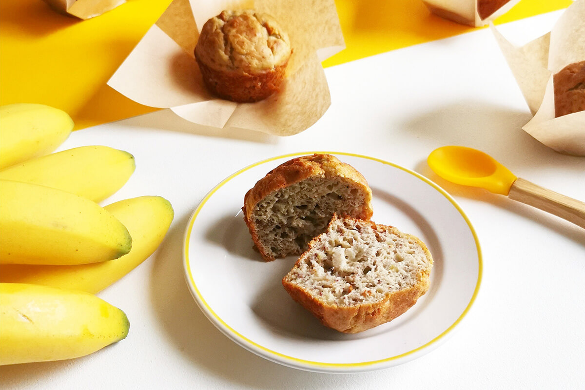 Baby banana muffins, one cut in half on a plate, the other on a paper cake case next to a bunch of bananas