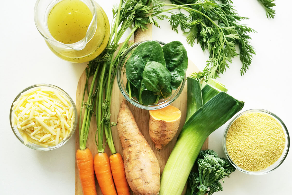 A chopping board with carrots, leek, sweet potato, spinach, broccoli, grated cheese, vegetable stock and uncooked couscous