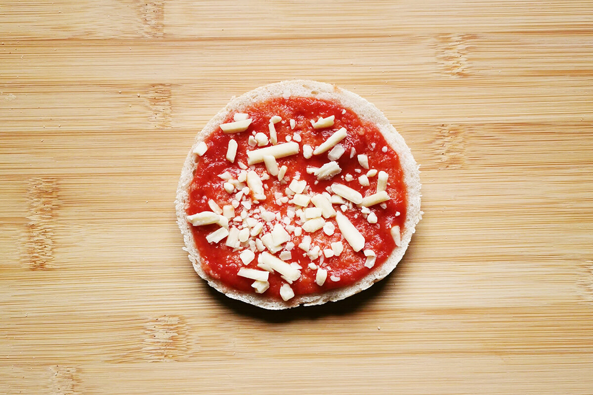 A halved English muffin, topped with pizza sauce and cheese, on a chopping board