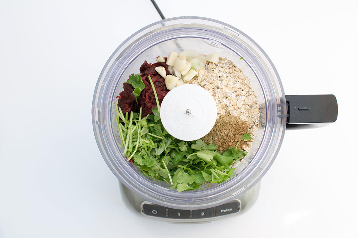 Grated beetroot in a blender with onion, garlic, oats, ground cumin and coriander, lemon juice, coriander sprigs and mint leaves