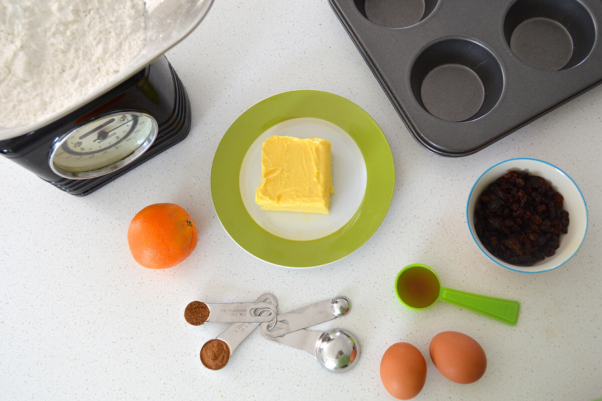 A surface with a plate of butter, a bowl of raisins, an orange, 2 eggs, a muffin case, some scales and some measuring spoons