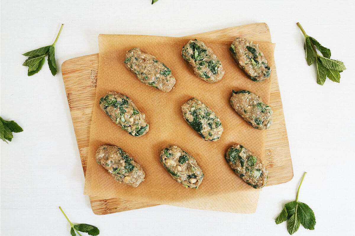 A chopping board with parchment paper and 9 uncooked lamb koftas on top