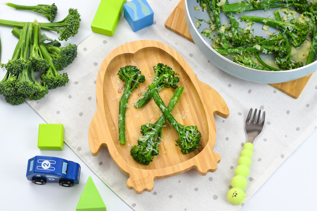 A serving of parmesan broccoli next to some tenderstem broccoli florets, a small bowl of Parmesan cheese and a garlic clove