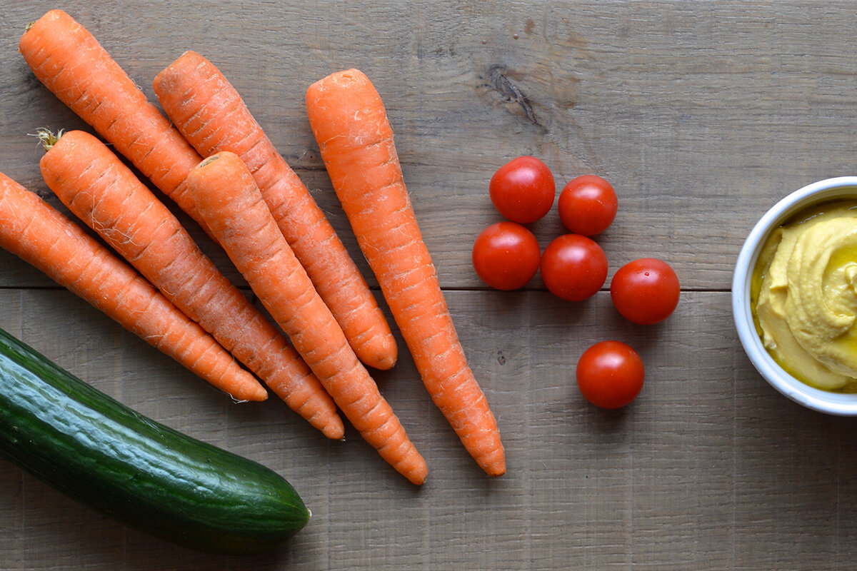 A table with a cucumber, carrots, cherry tomatoes and a bowl of hummus