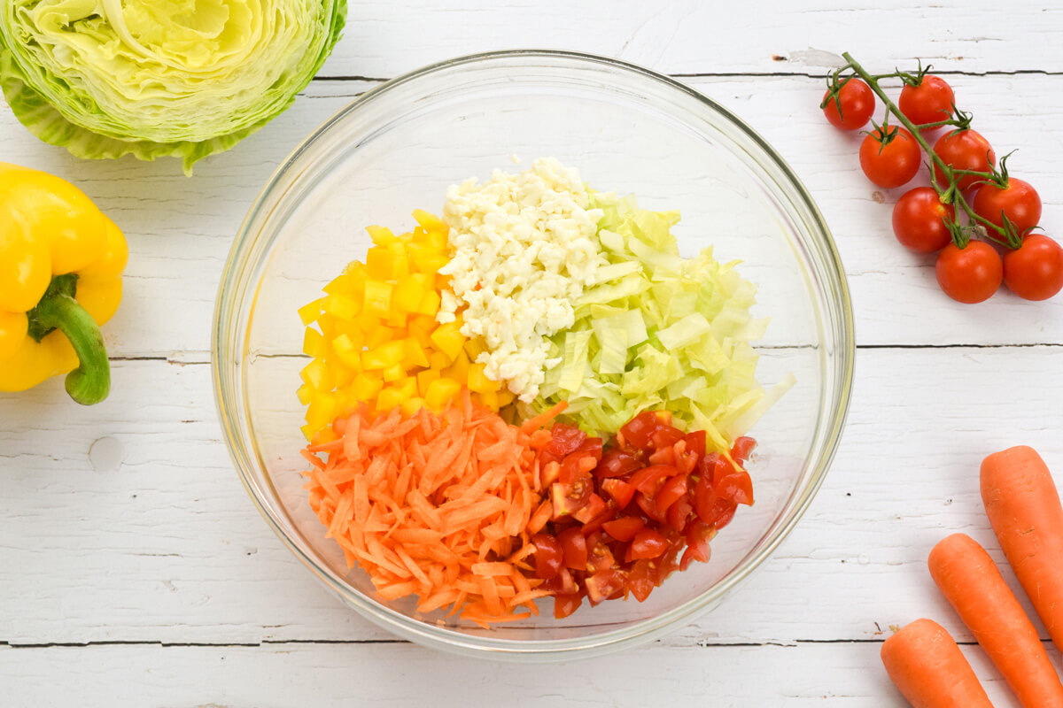 A large glass bowl of rainbow salad (lettuce, pepper, carrot, tomato and mozzarella) next to some carrots, lettuce and cherry tomatoes