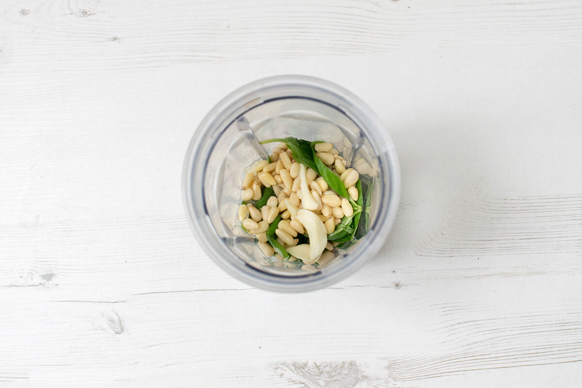 A blender with basil leaves, pine nuts, garlic and lemon juice in it