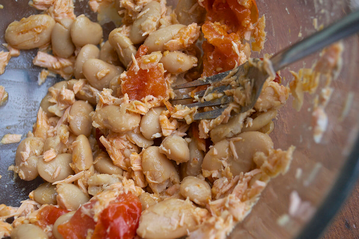 A glass bowl with butter beans, cherry tomatoes and salmon being mashed together with a fork