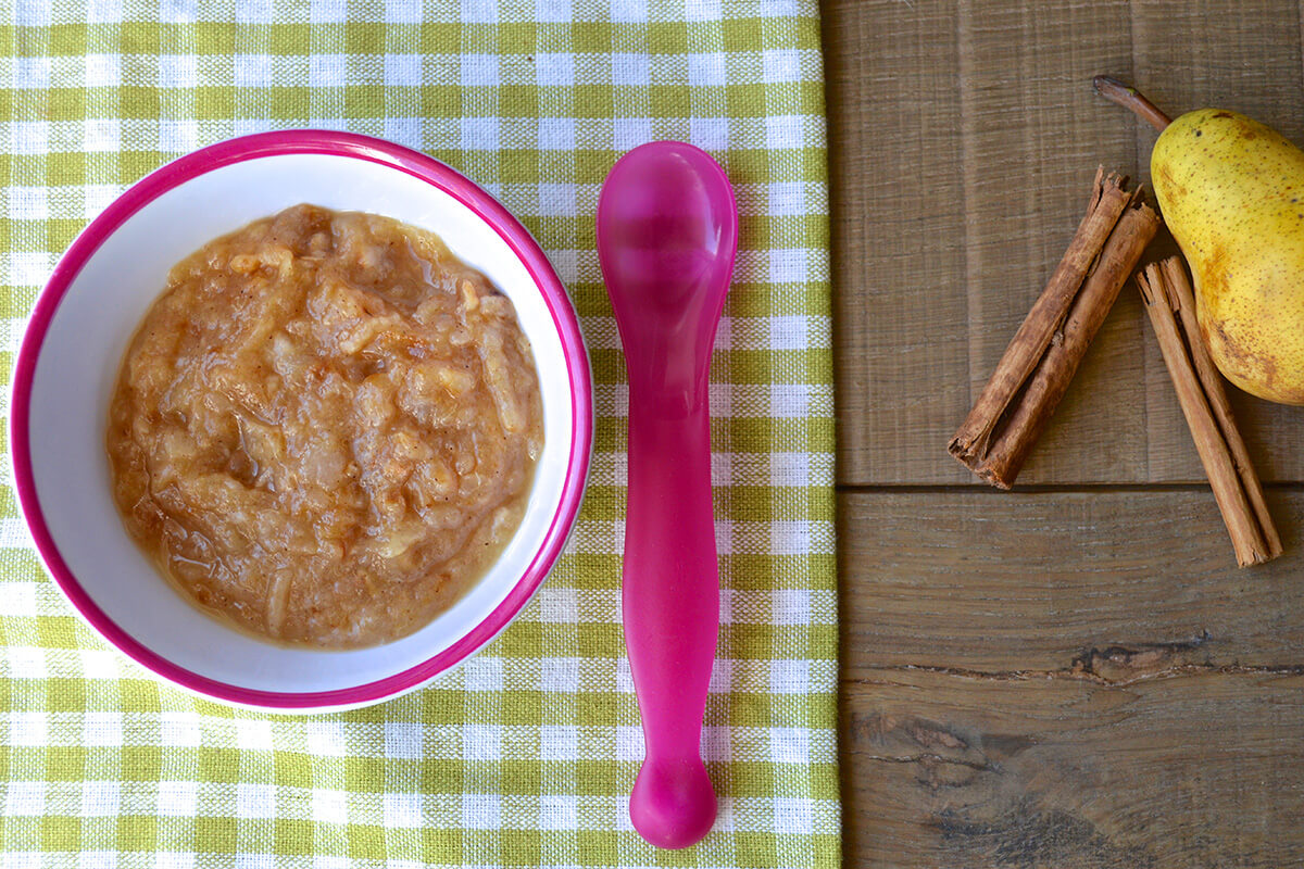 A bowl of spiced apple puree next to a whole pear and two cinnamon sticks