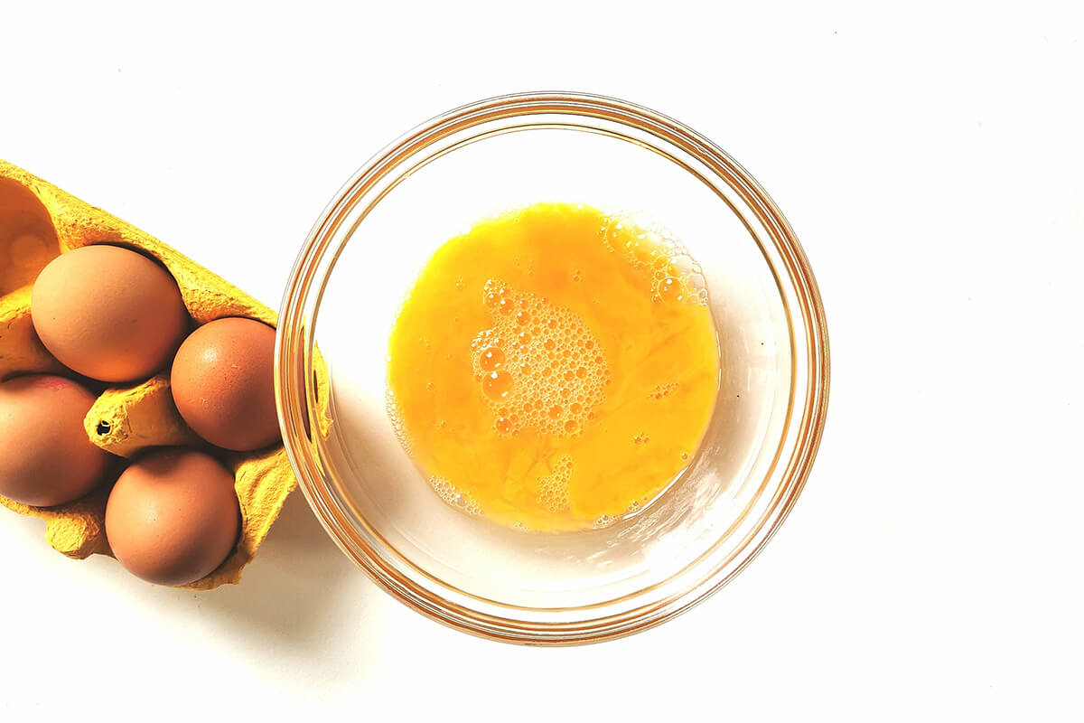 A bowl of whisked eggs next to a crate of 4 eggs