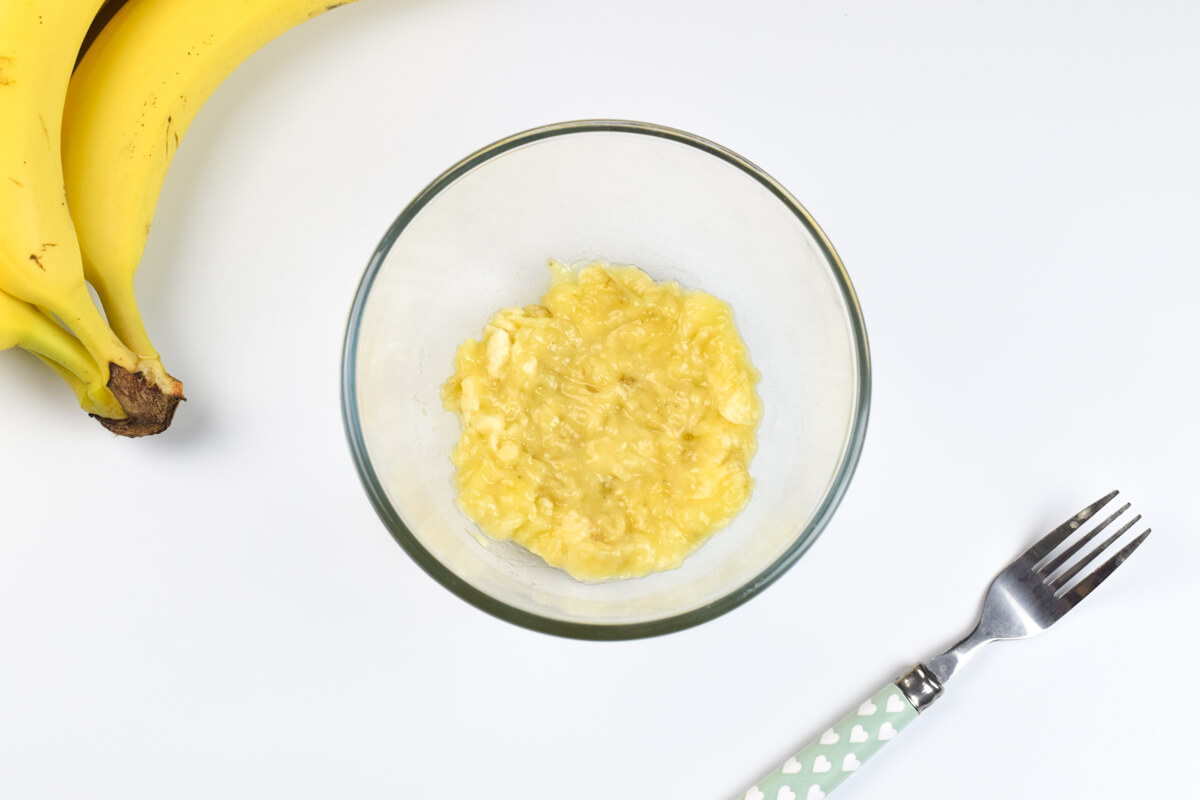 A glass bowl with mashed banana next to a bunch of bananas